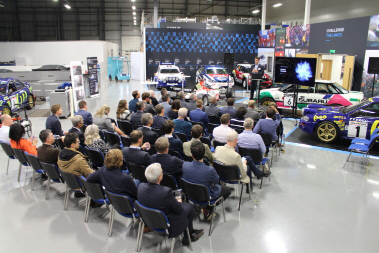 Photo of the challenge from previous years: a group of people sat in rows, with a presenter on stage, surrounded by vehicles at the Prodrive offices